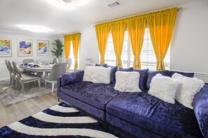 A living room with a purple sofa, yellow curtains, and a dining area with a table and chairs.