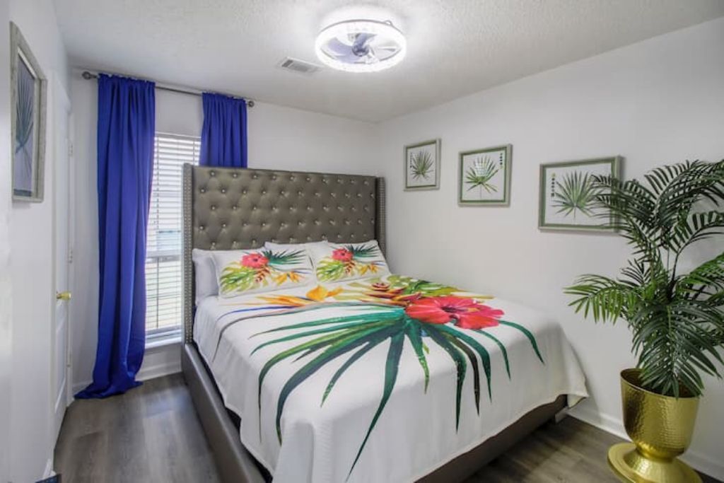 A bedroom with a tufted headboard, a bed with a floral bedspread, a potted plant, and framed botanical prints on the wall.