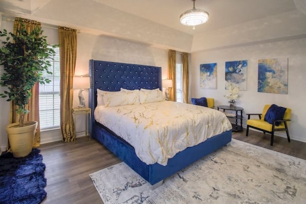 Une chambre à coucher avec un grand lit, une table de chevet, une lampe, une chaise, une petite table et une plante en pot.
