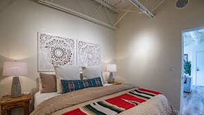 A bedroom with a large bed, two bedside lamps, decorative wall panels, and a view into another room. - Chic View Loft at Historic Plaza - Spacious 470 Sq Ft (Santa Fe)