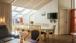Un comedor con sillas de madera y una mesa, un televisor montado en la pared y vistas del exterior a través de un tragaluz.
