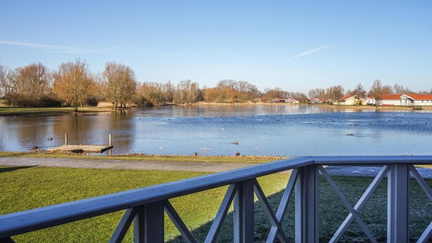 Vista de un lago con un muelle de madera, orillas cubiertas de césped y casas al fondo.