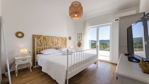 Une chambre avec un lit blanc, une table de chevet avec une lampe, une grande fenêtre donnant sur un beau paysage et un téléviseur posé sur un meuble.