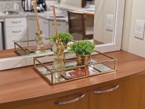 A kitchen counter with a mirror reflecting a stove, a sink, and cabinets. There are decorative items including a glass bottle with a gold ribbon, a small potted plant, and a framed picture.