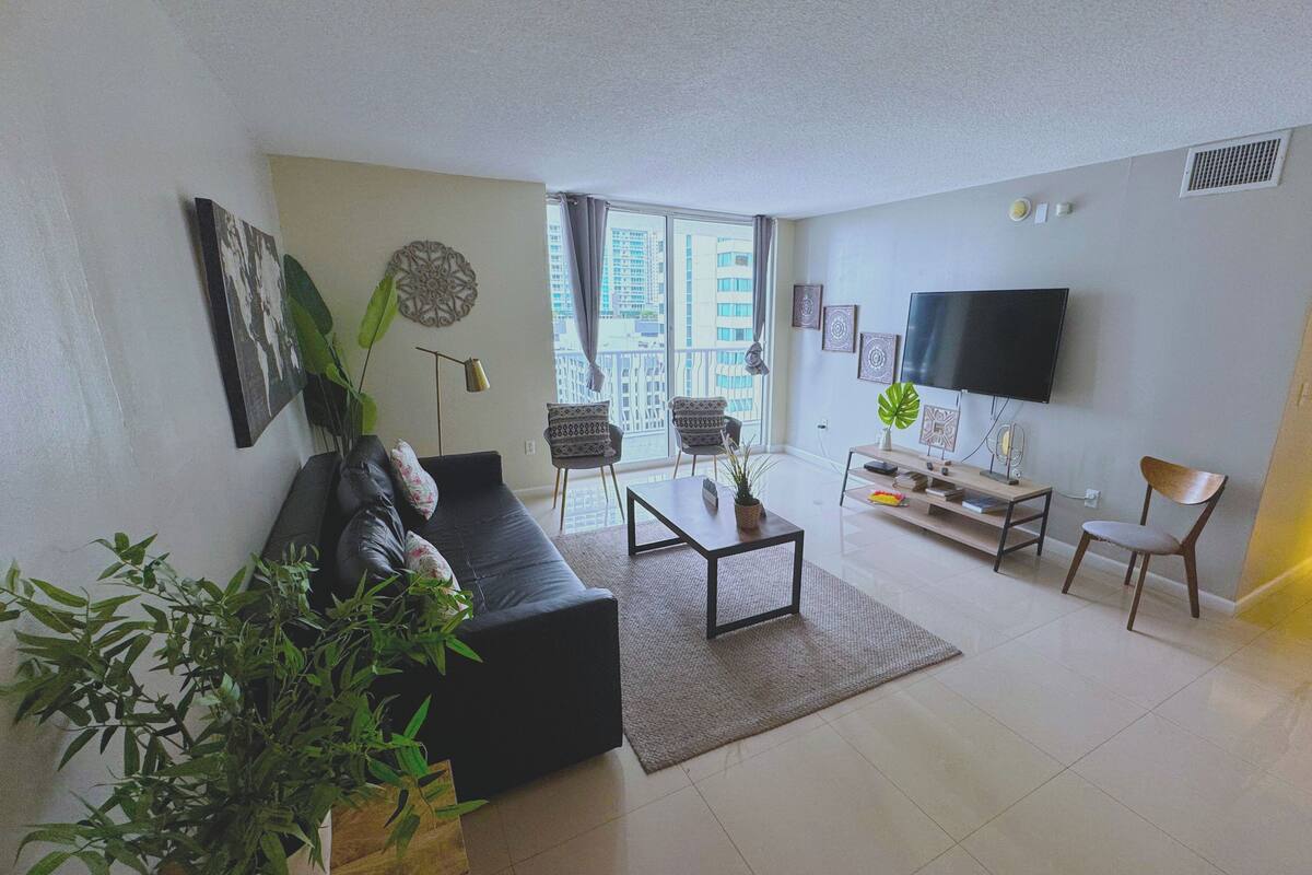 Uma sala de estar moderna com um sofá preto, uma mesa de centro, uma televisão e uma janela grande com vista para a cidade.