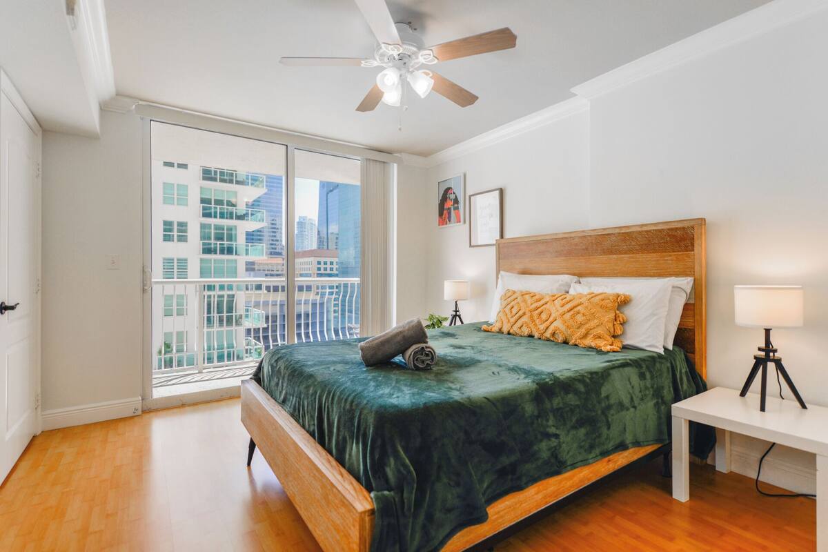 Quarto com cama grande, ventilador de teto, mesa de cabeceira e vista da cidade através de uma janela ampla.