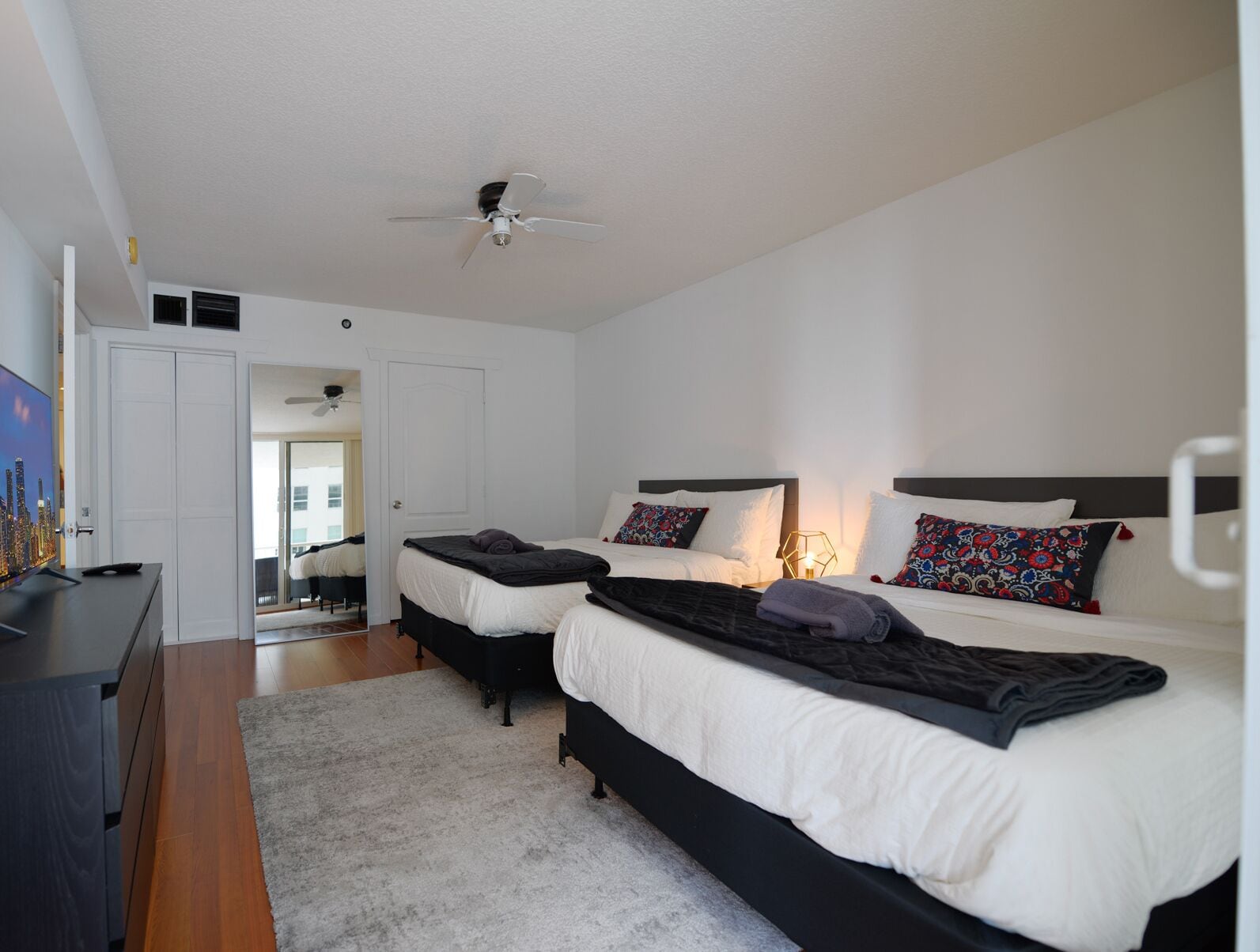 A bedroom with two beds, a ceiling fan, and a view of a cityscape through a window.