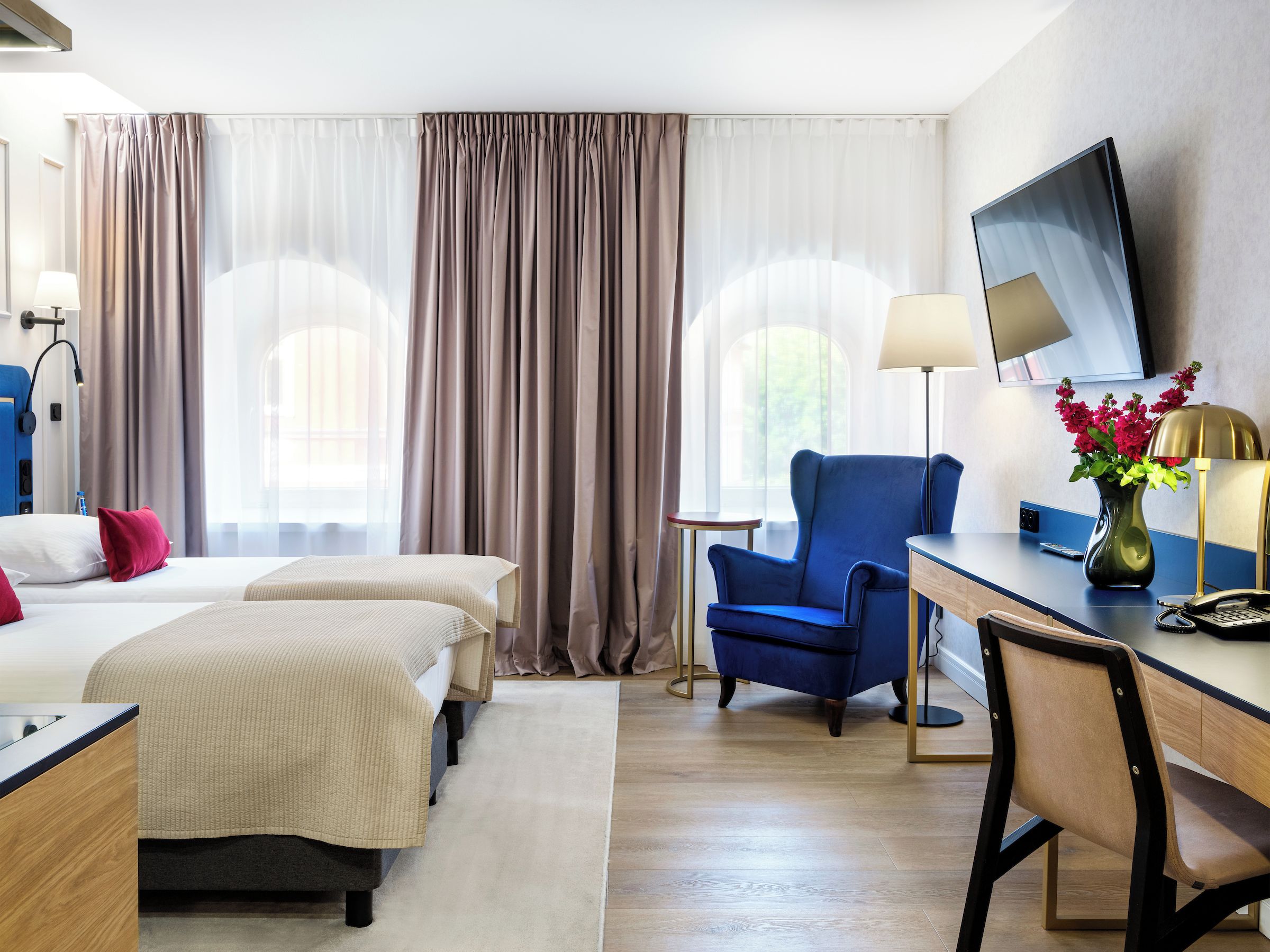 A hotel room with two beds, a desk, a chair, a TV, and a vase with flowers.