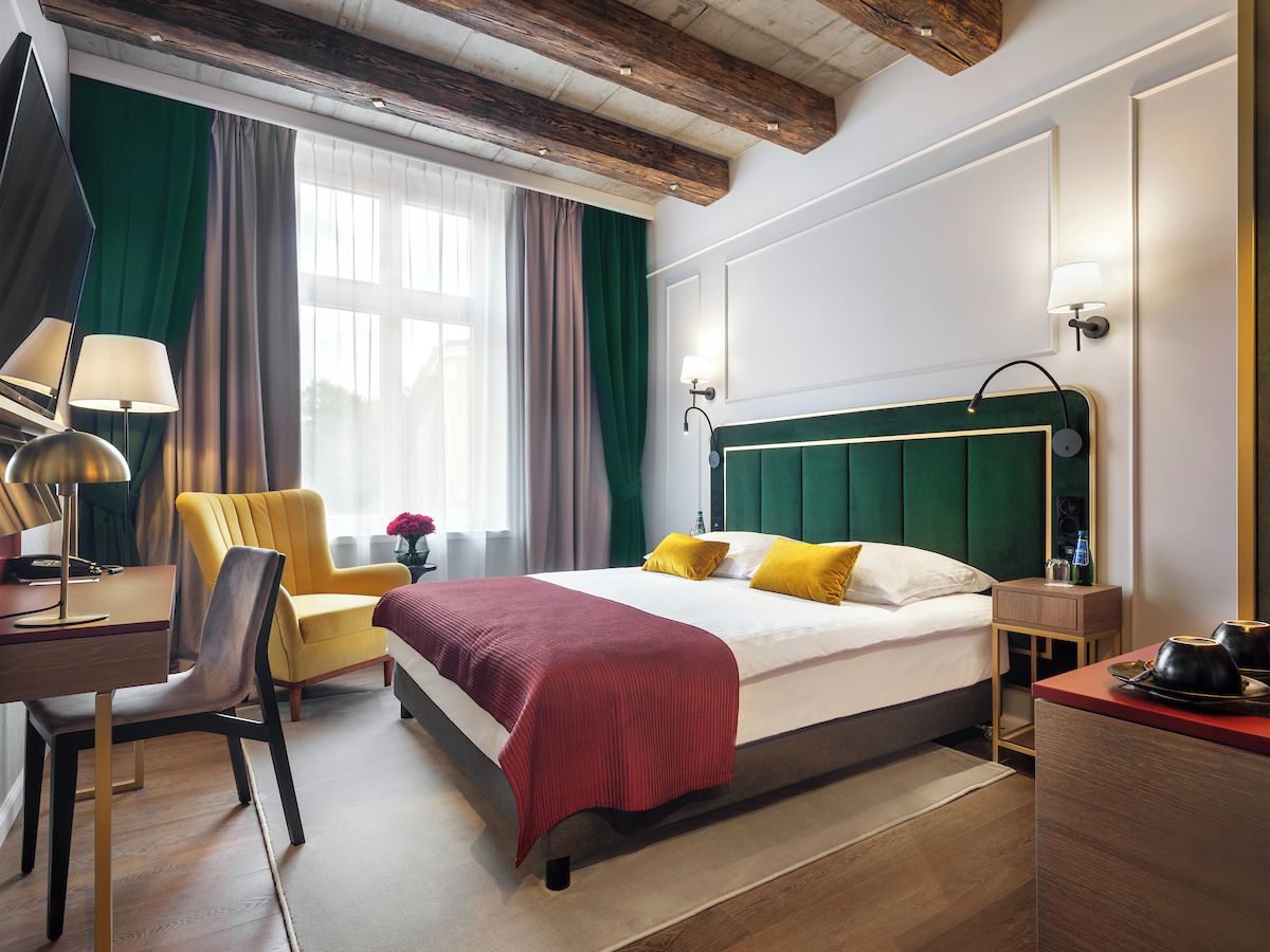 Ein Hotelzimmer mit Bett, Schreibtisch, Stuhl, Nachttisch, Lampe und einem Fenster mit Vorhängen.