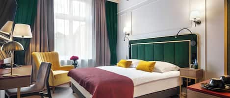 A hotel room with a bed, a desk, a chair, a bedside table, a lamp, and a window with curtains.