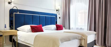 Habitación de hotel con una cama grande, dos mesitas de noche, una lámpara, una botella de agua y una ventana con cortinas.