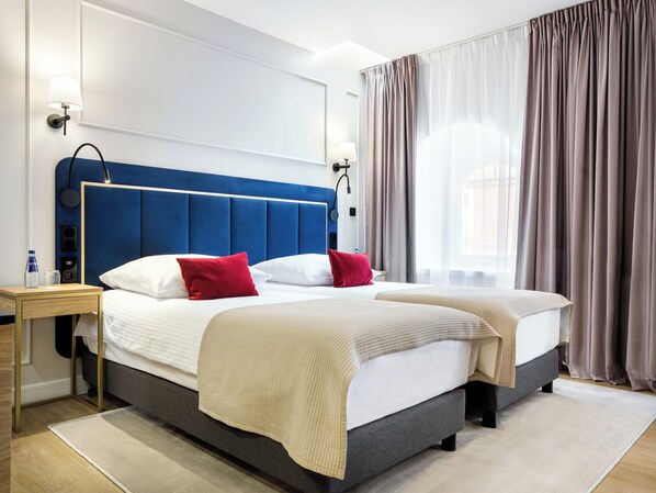 A hotel room with a large bed, two bedside tables, a nightstand, a lamp, a water bottle, and a window with curtains. - Hotel Golden Queen (Kraków)
