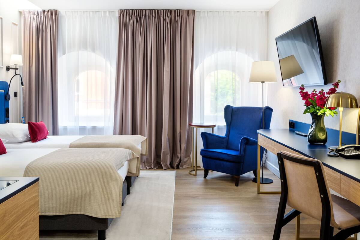 A hotel room with two beds, a desk, a chair, a TV, and a vase with flowers.
