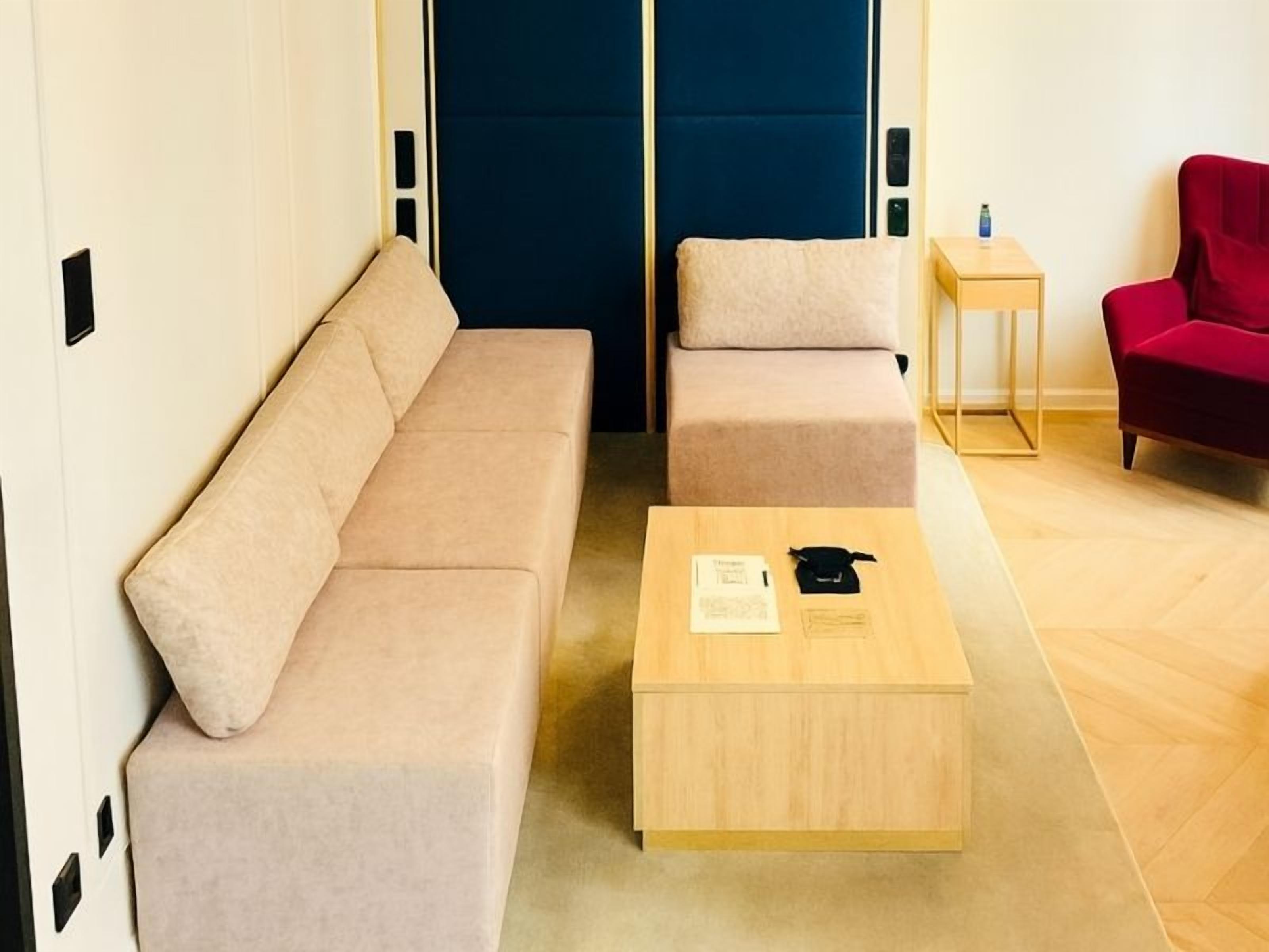 Een moderne hotelkamer met een beige bank, een kleinere bank, een houten salontafel en een rode fauteuil.