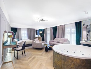 Una habitación de hotel moderna con una cama grande, dos sillones, un sofá, una bañera de hidromasaje redonda, un televisor y un espejo.