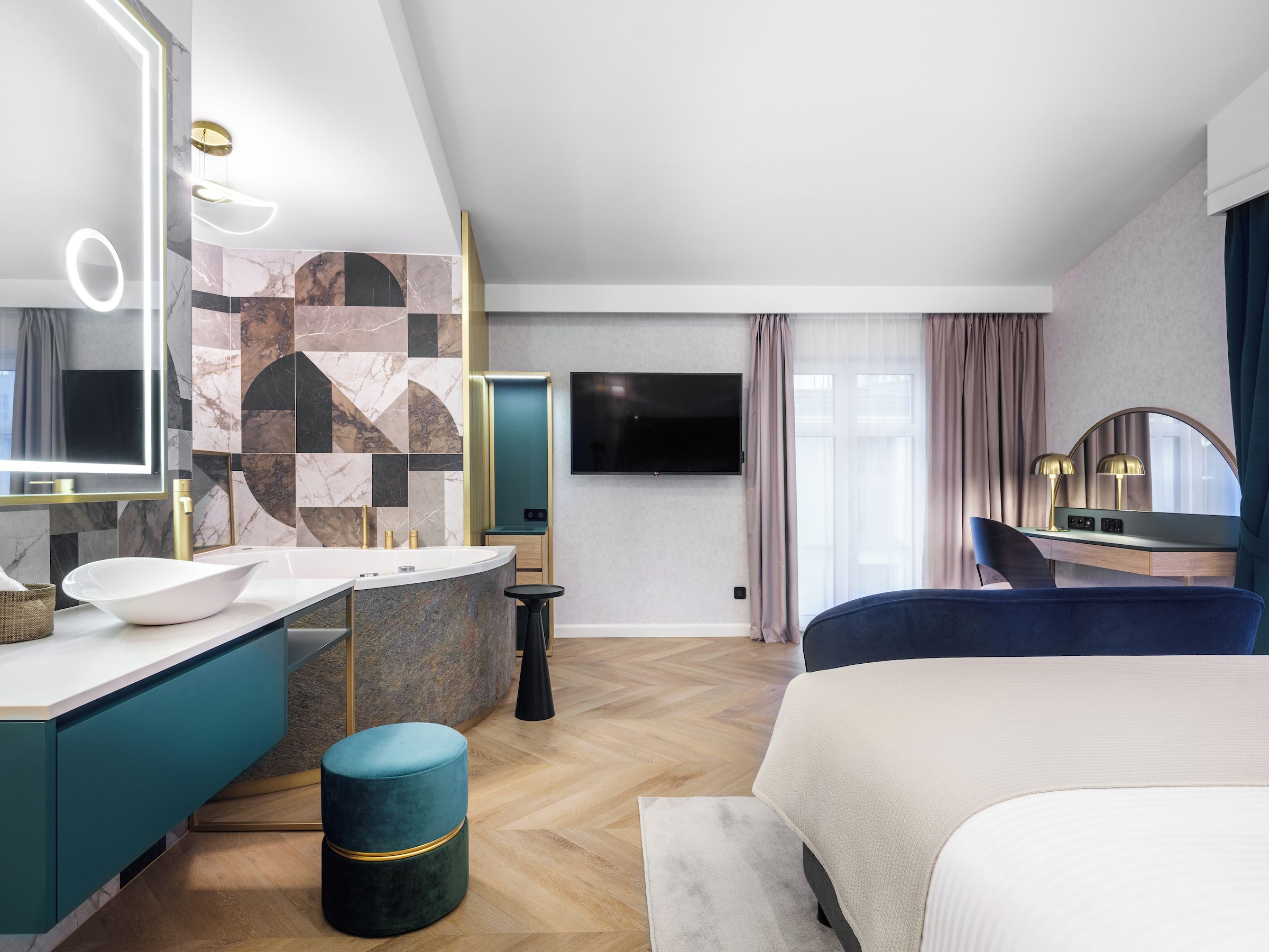 A modern hotel room with a large bed, a vanity with a mirror, a television, and a seating area with a table and chairs.