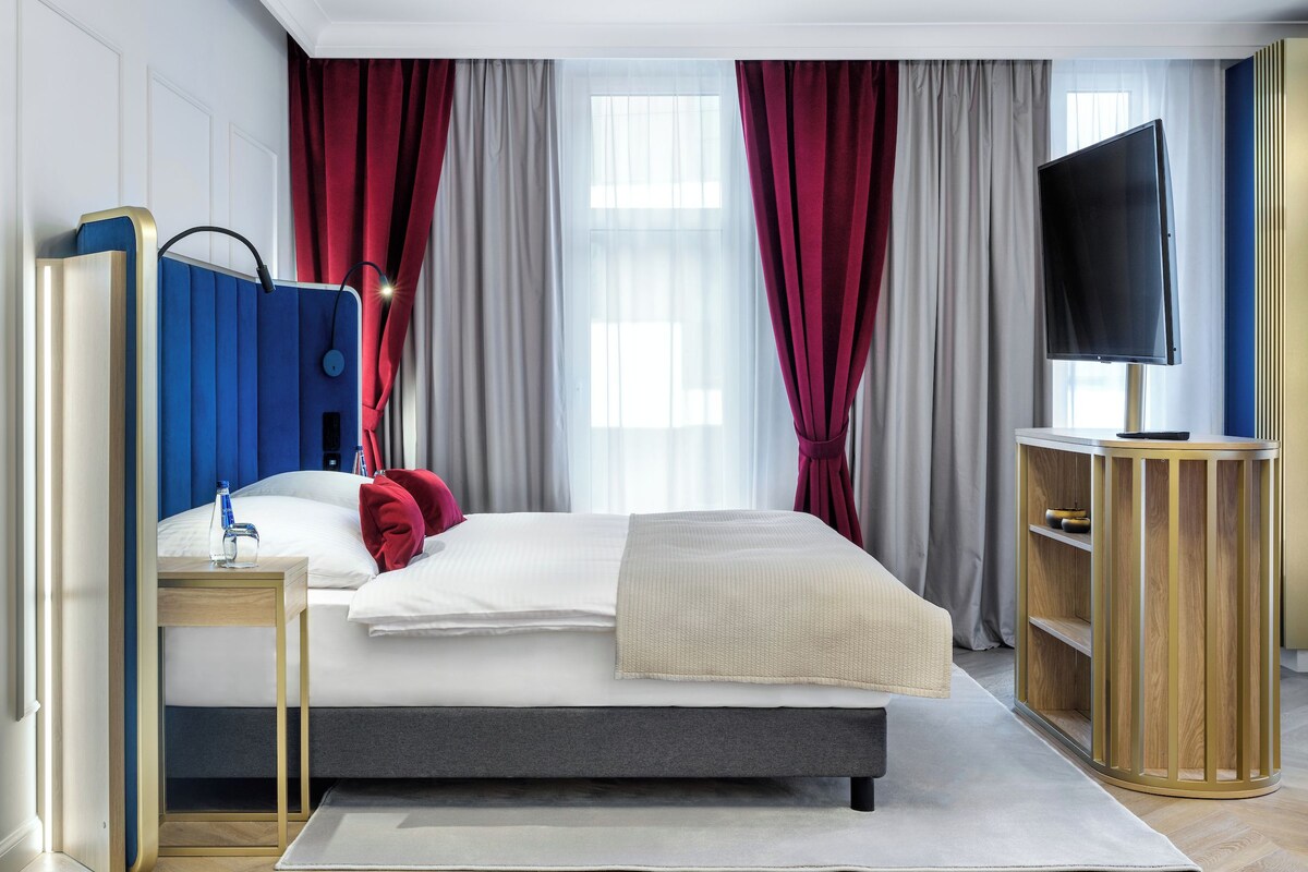 A modern hotel room with a bed, bedside tables, a TV, and a wardrobe.