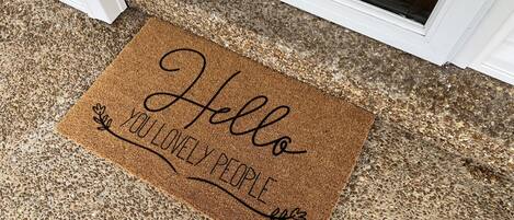 A brown doormat with the text "Hello YOU LOVELY PEOPLE" placed at a door entrance.