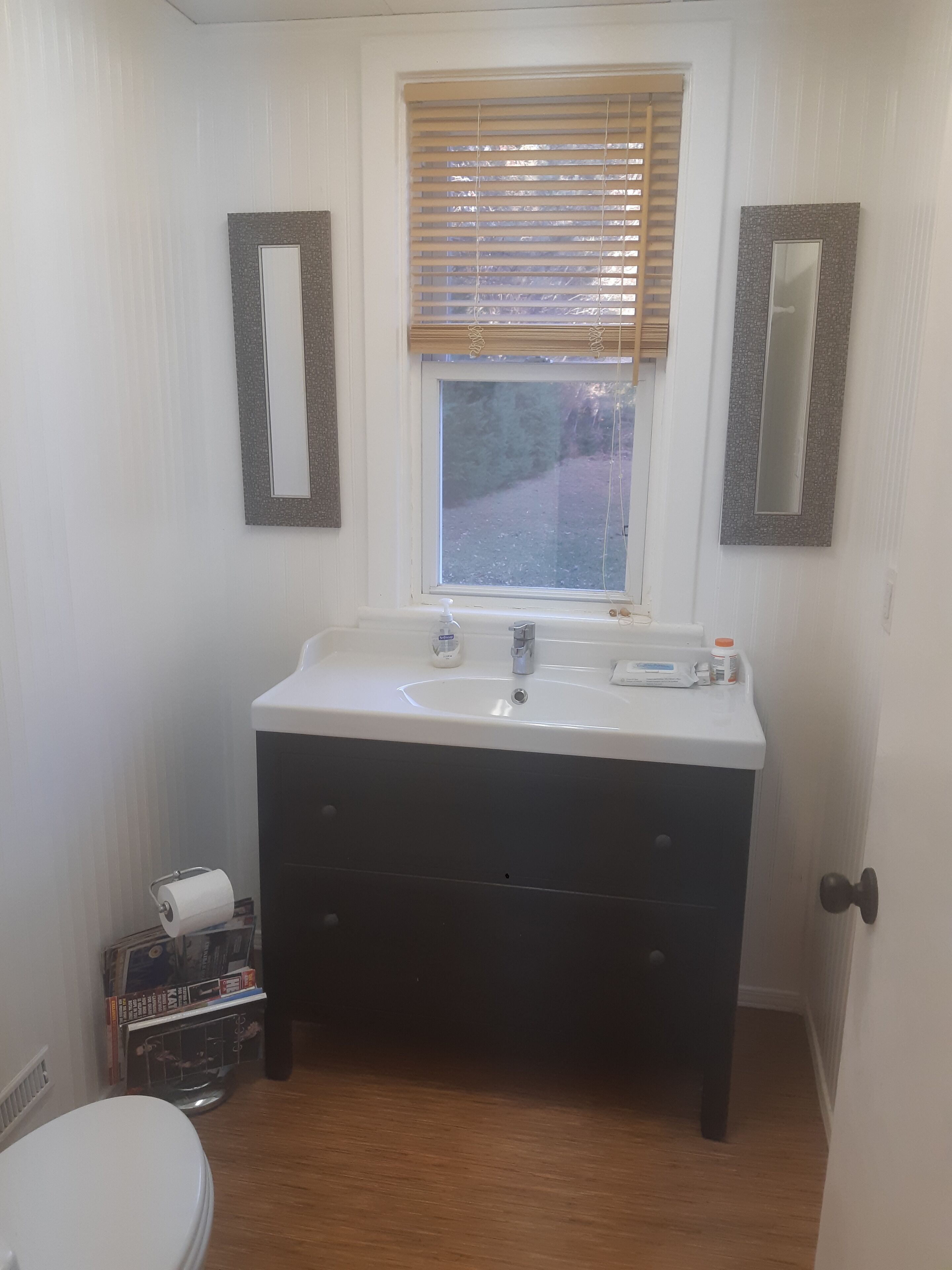 A bathroom with a white sink, dark vanity, two wall mirrors, a window with blinds, and a toilet with a roll of toilet paper.