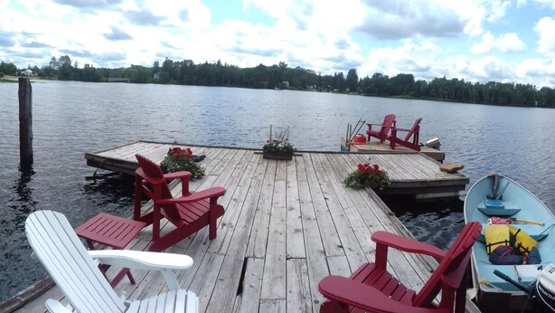 A wooden dock with red Adirondack chairs and a small boat.