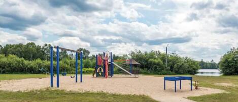 A playground with a slide and climbing structures, a blue table, and a path leading to a body of water.