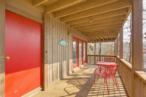 Una terraza de madera con una puerta roja, un letrero con forma de pez y una mesa con sillas rojas.