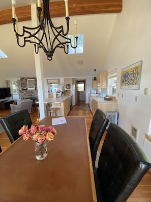 A dining area with a wooden table, black chairs, and a vase of flowers. There is a kitchen in the background with white cabinets and a window.