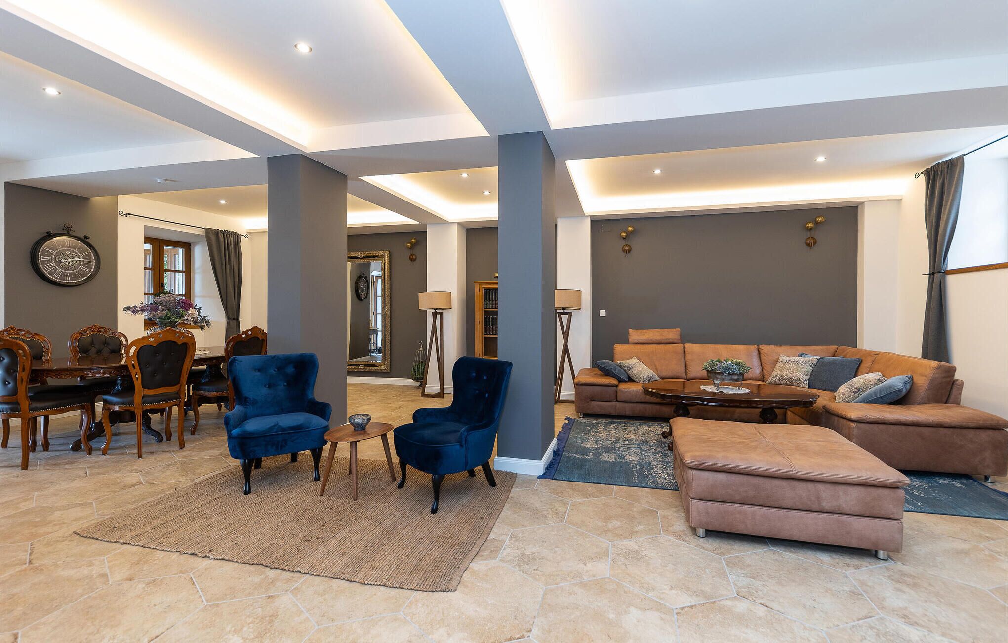 A spacious living room with a brown leather sofa, blue armchairs, and a dining area with a wooden table and chairs.