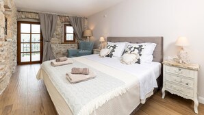 Een slaapkamer met een stenen muur, een groot bed met veel kussens, een nachtkastje en een raam met gordijnen.