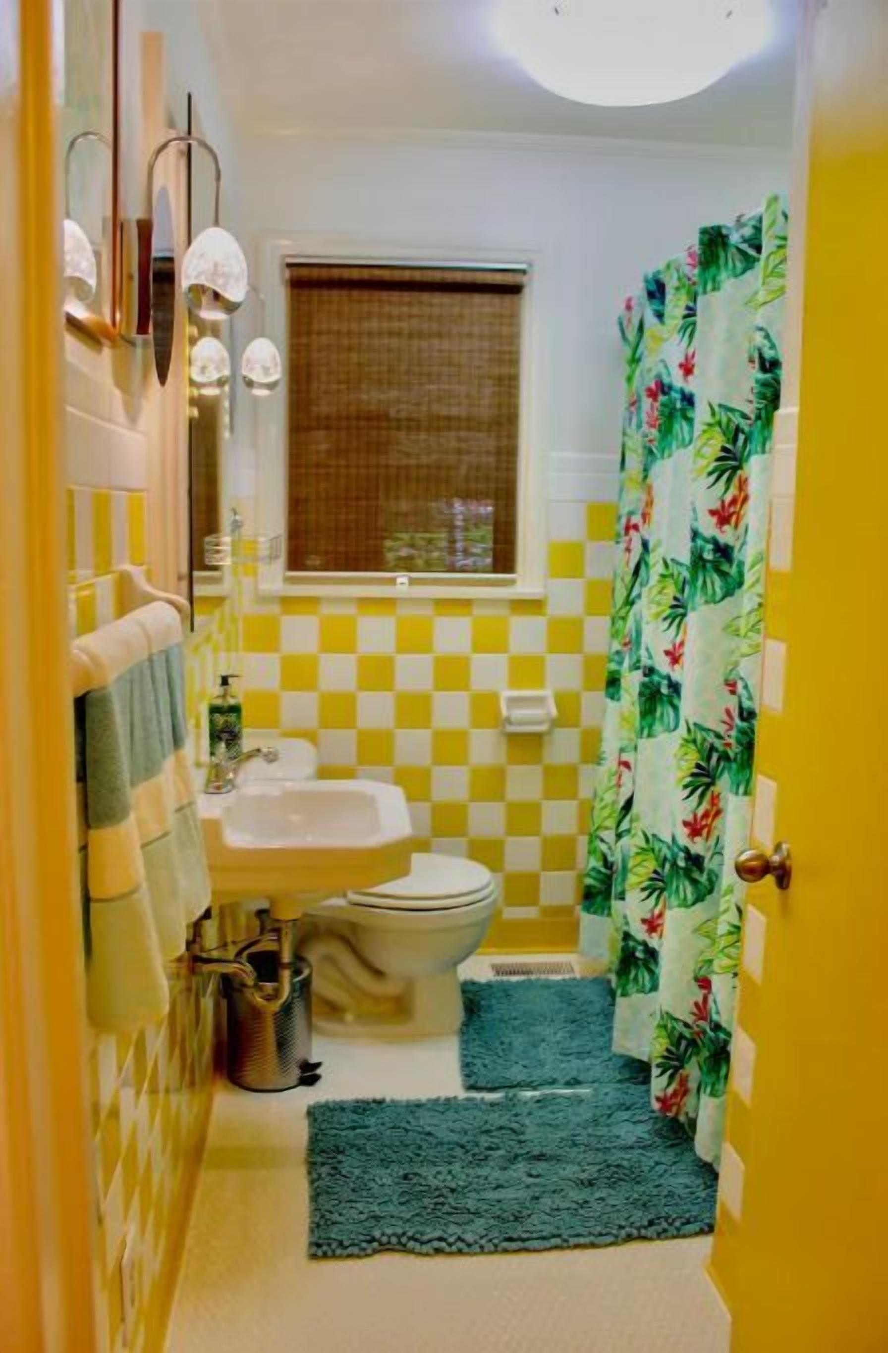 Un baño con pared a cuadros amarillo y blanco, un inodoro, un lavamanos y una cortina de ducha con motivos tropicales.