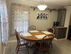 A dining area with a wooden table, four chairs, floral curtains, a wall-mounted painting of cows, and a kitchen area with a coffee maker.