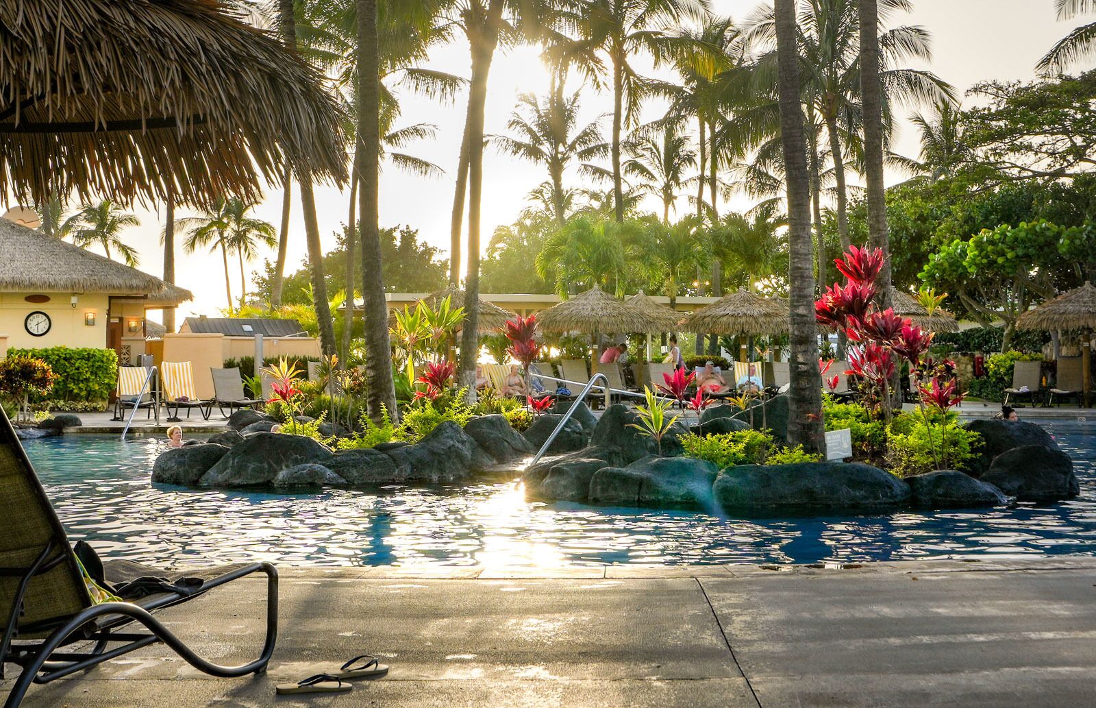 Un espace piscine aménagé dans un complexe hôtelier, avec des chaises longues, un bâtiment à toit de chaume et une végétation tropicale.