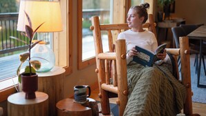 A person seated in a wooden chair reading a book, with a lamp and a potted plant on a wooden table beside them. - Modern Chalet with Lake Views in Marina Area (Mossyrock)