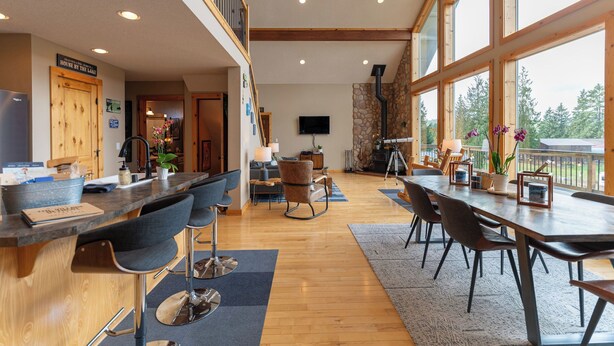 A spacious living area with a high ceiling, a stone fireplace, and a large window offering a view of the outdoors. - Modern Chalet with Lake Views in Marina Area (Mossyrock)