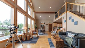 A spacious living room with a telescope, wooden flooring, a leather sofa, and a staircase leading to an upper level. - Modern Chalet with Lake Views in Marina Area (Mossyrock)