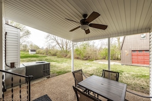 A covered patio with a table and chairs, an air conditioning unit, and a red garage door in the background.