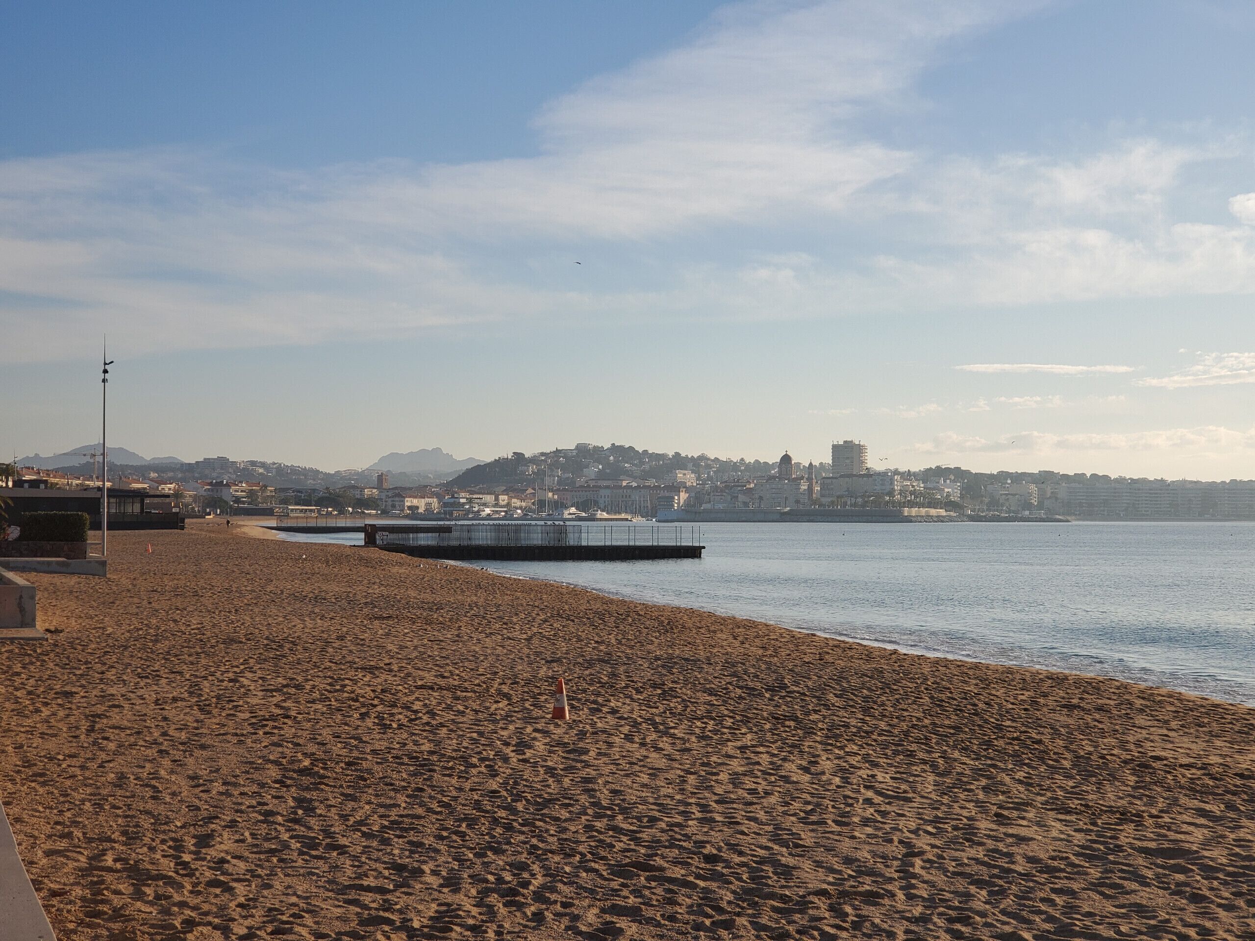 A beach with a pier extending into calm waters, a distant cityscape, and a clear sky.