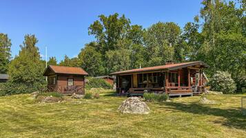 A rustic cabin with a porch and a smaller adjacent building, surrounded by a grassy area and trees.