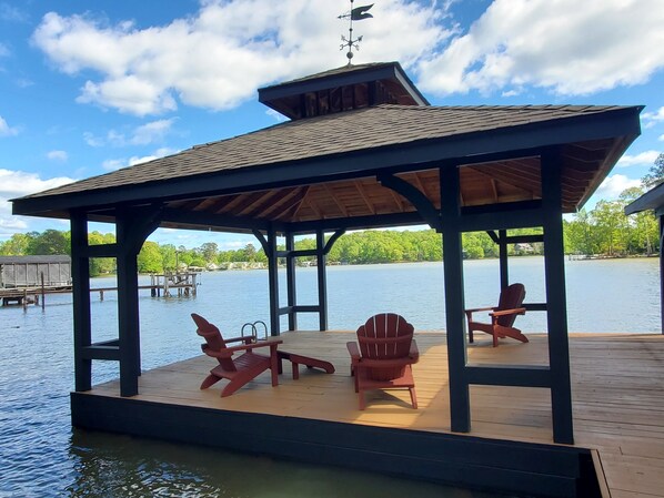 A wooden deck with a gazebo, two Adirondack chairs, and a dock extending into a lake.