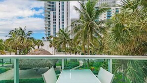 A balcony with a glass table and chairs, overlooking a landscaped garden with palm trees and a building in the background. - Beautiful Vacation Rental Directly on beach (Hollywood)