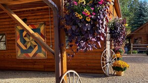 Un bâtiment en bois orné d’un motif géométrique sur le mur, un chariot en bois et des pots de fleurs posés sur le sol.