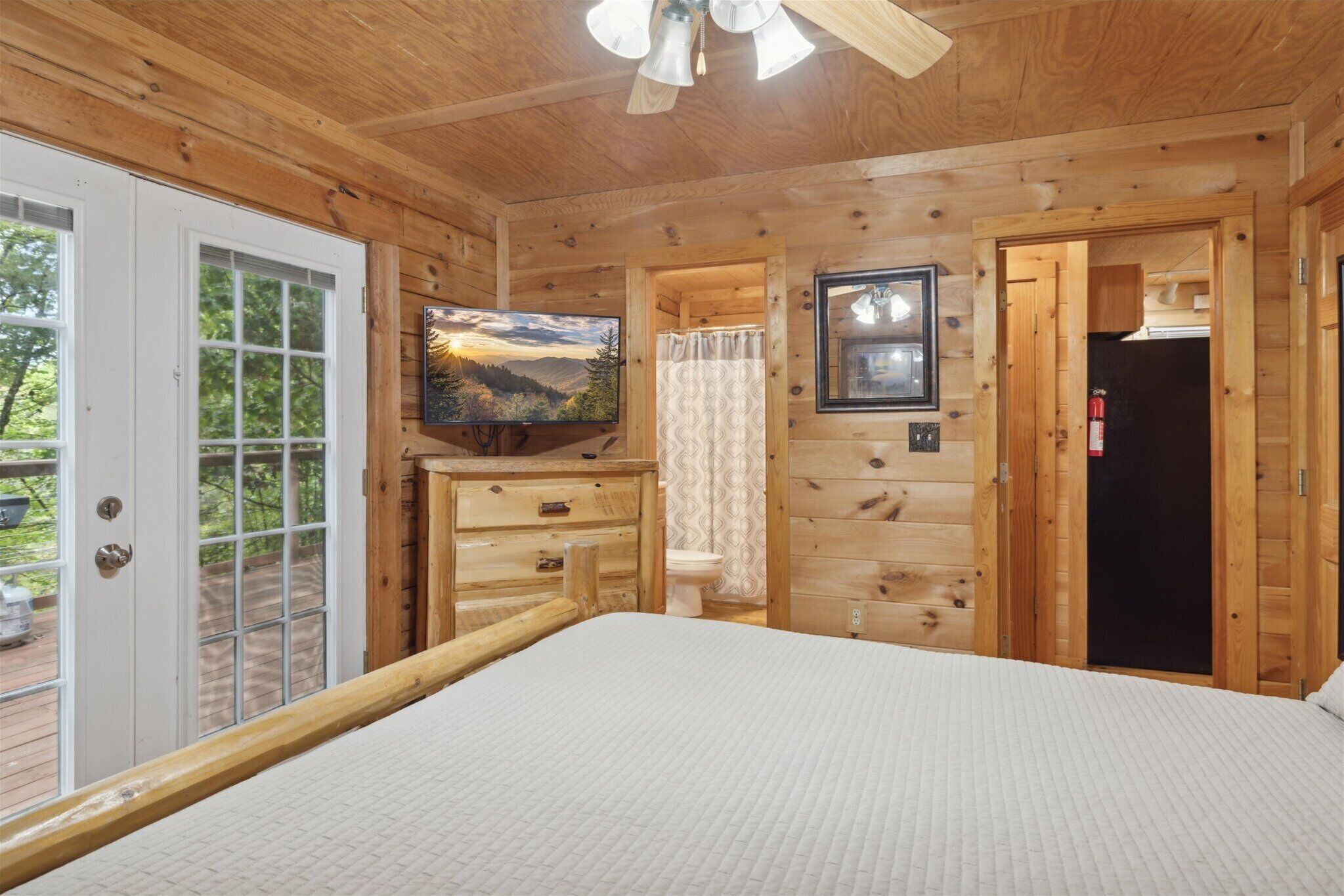 Un dormitorio de cabaña de madera con una cama, un televisor, una ventana con vistas panorámicas y una puerta que da a un baño.