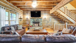 A log cabin interior with a stone fireplace, leather sofas, and a wooden staircase. - Hot Tub & Fire Pit: Getaway Near Nantahala Gorge! (Franklin)