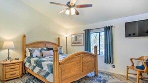 A bedroom with a wooden bed, a nightstand, a lamp, a ceiling fan, a TV, and a window with curtains. - Sunny Tucson Home w/ Patios on 5 Acres! (Tucson)