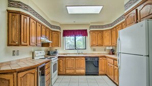 A kitchen with wooden cabinets, a refrigerator, a dishwasher, and a window with a red valance. - Sunny Tucson Home w/ Patios on 5 Acres! (Tucson)