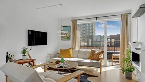 Een moderne woonkamer met een bank, fauteuil en salontafel, een flatscreen-tv en een groot raam met uitzicht op de stad.
