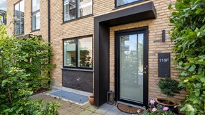 Een modern gebouw van baksteen met een zwarte deur en ramen, een kleine tuin met planten en bloemen, en een huisnummerplaatje.