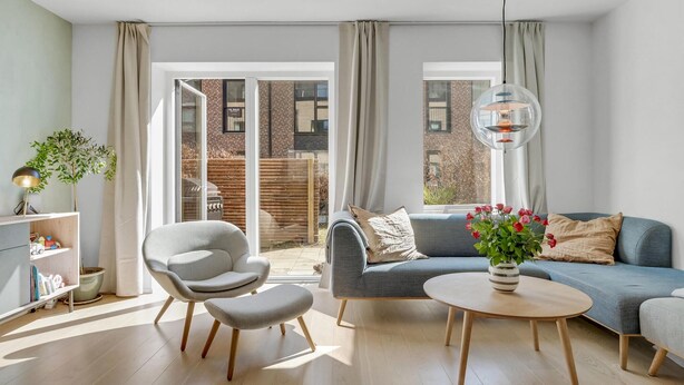 Een moderne woonkamer met een grijze bank, een ronde salontafel en een grijze fauteuil. Er is een boekenkast met boeken en een kleine plant. De kamer heeft grote ramen met gordijnen en uitzicht op een gebouw van baksteen.