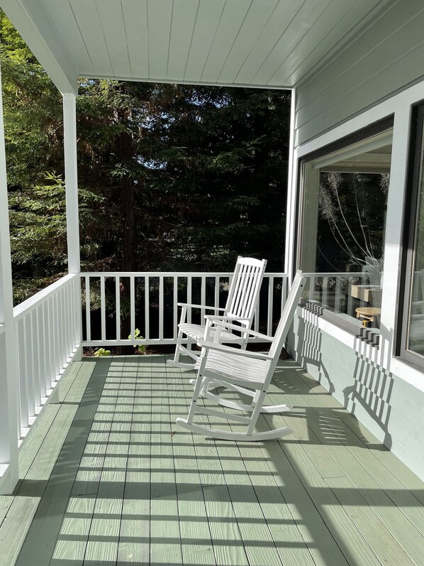 A covered porch with a white railing, a rocking chair, and a window overlooking a forested area.