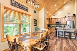 A rustic interior with a dining area, wooden chairs, a kitchen with wooden cabinets, and a sign above the windows.